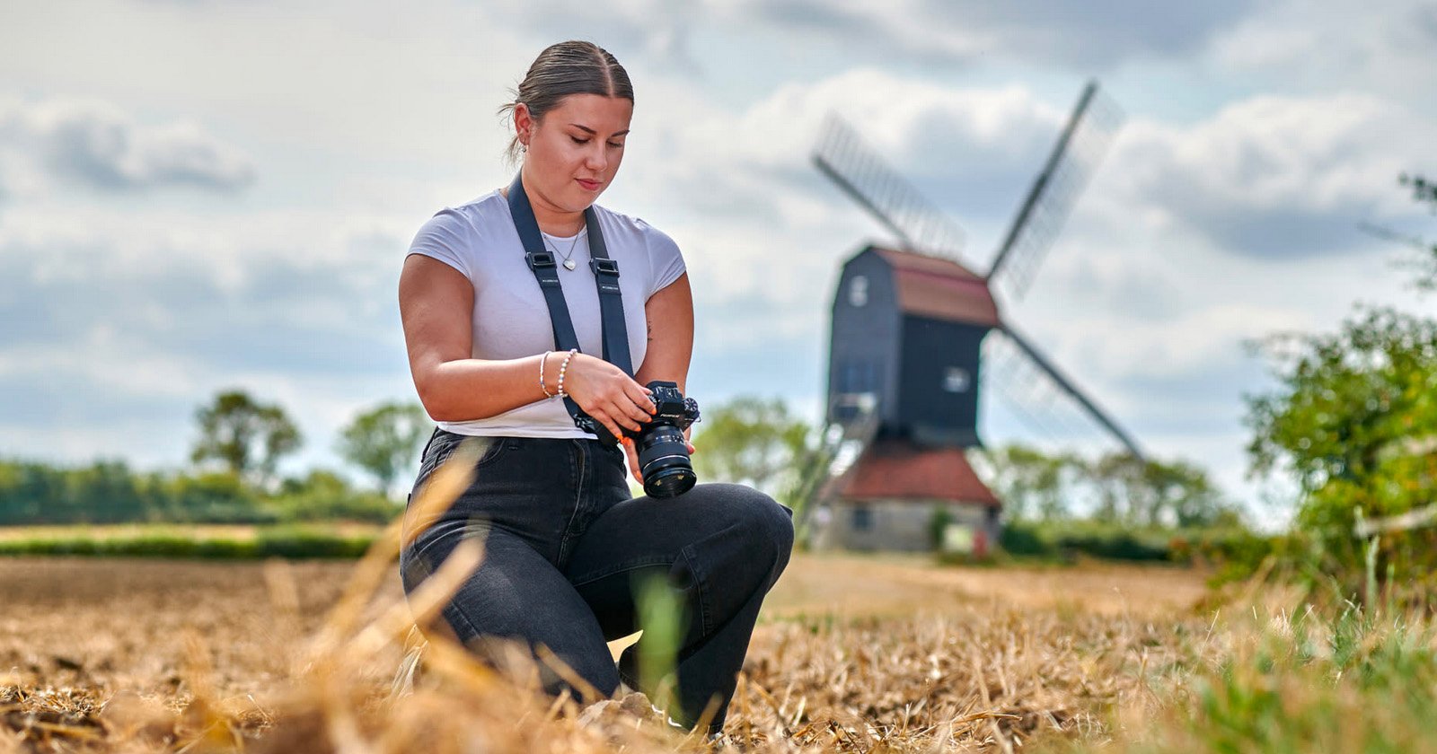 A woman kneels in a dry field, looking down at her camera. She wears a white shirt and black pants. A windmill with a red roof stands in the background under a partly cloudy sky.