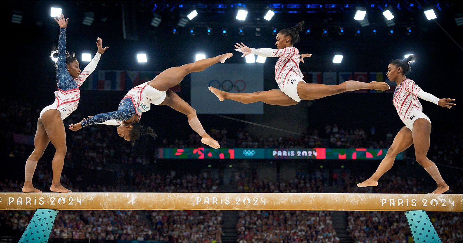 Four gymnasts in mid-routine perform dynamic moves on a balance beam labeled Paris 2024, with an audience and large screen displaying Paris 2024 in the background. The gymnasts wear red, white, and blue leotards in a stadium lit with spotlights.
