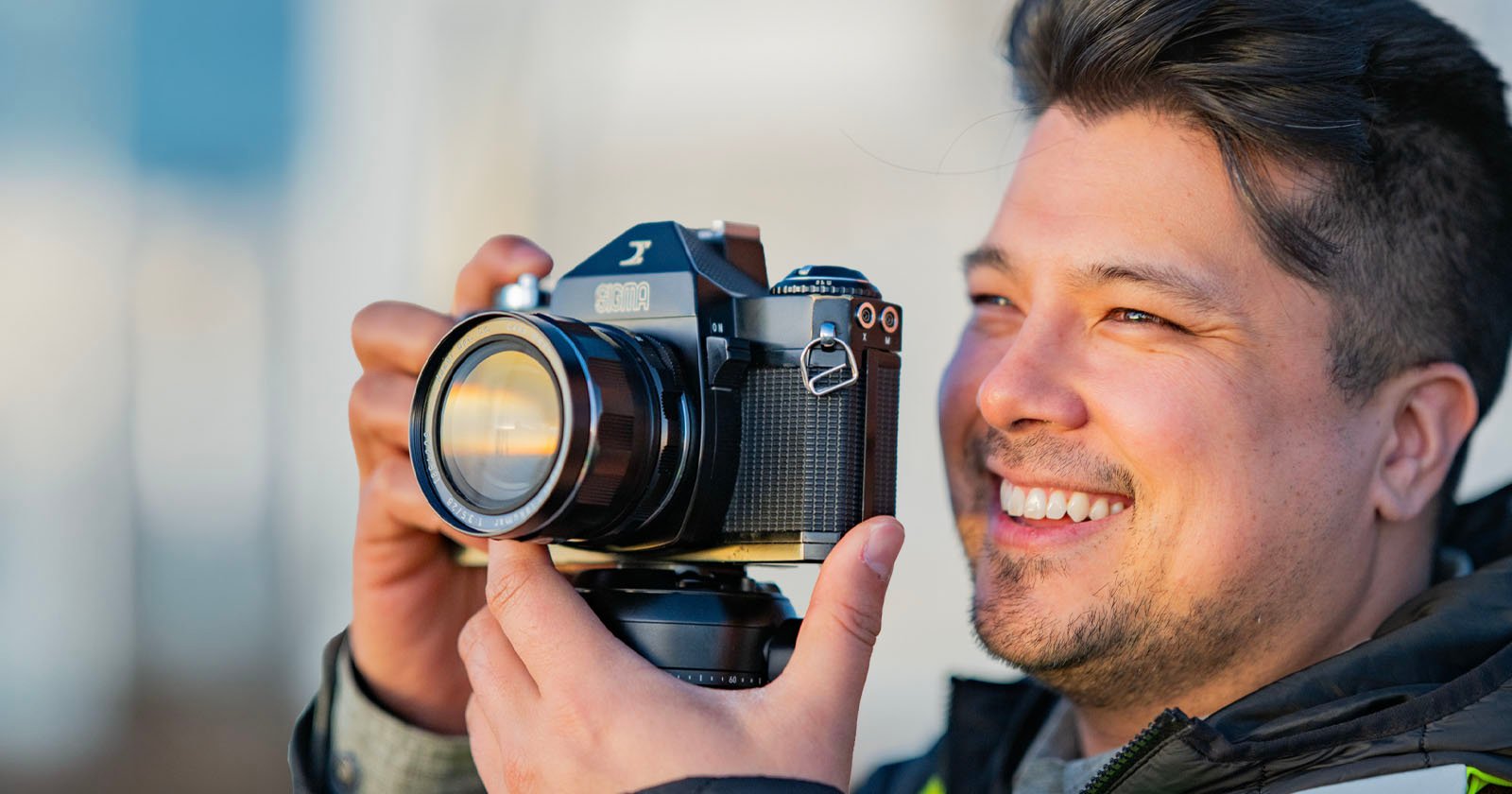 A man smiles while holding a vintage film camera up to his face, ready to take a photo outdoors in natural light.