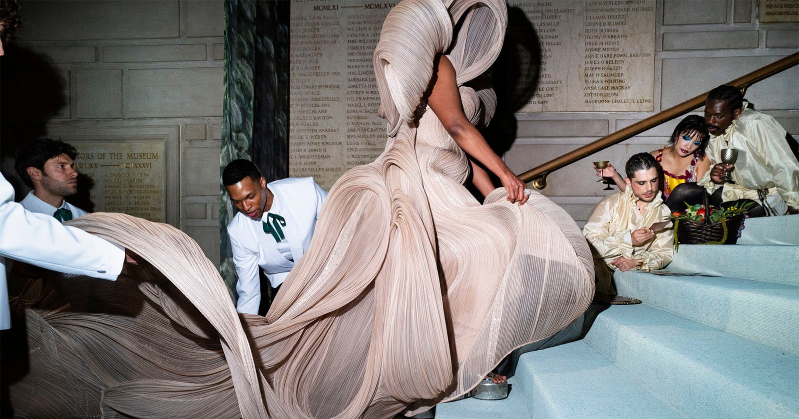 A person in an elaborate, flowing beige gown walks up stairs while attendants in white help with the dress. Several people in gold outfits sit on the steps, some eating and chatting, against a stone wall with engraved text.