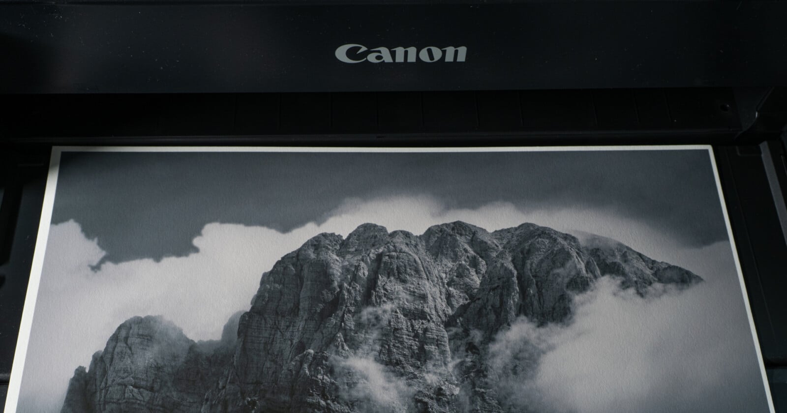 A black-and-white photograph of a mountain surrounded by clouds is being printed from a Canon printer.