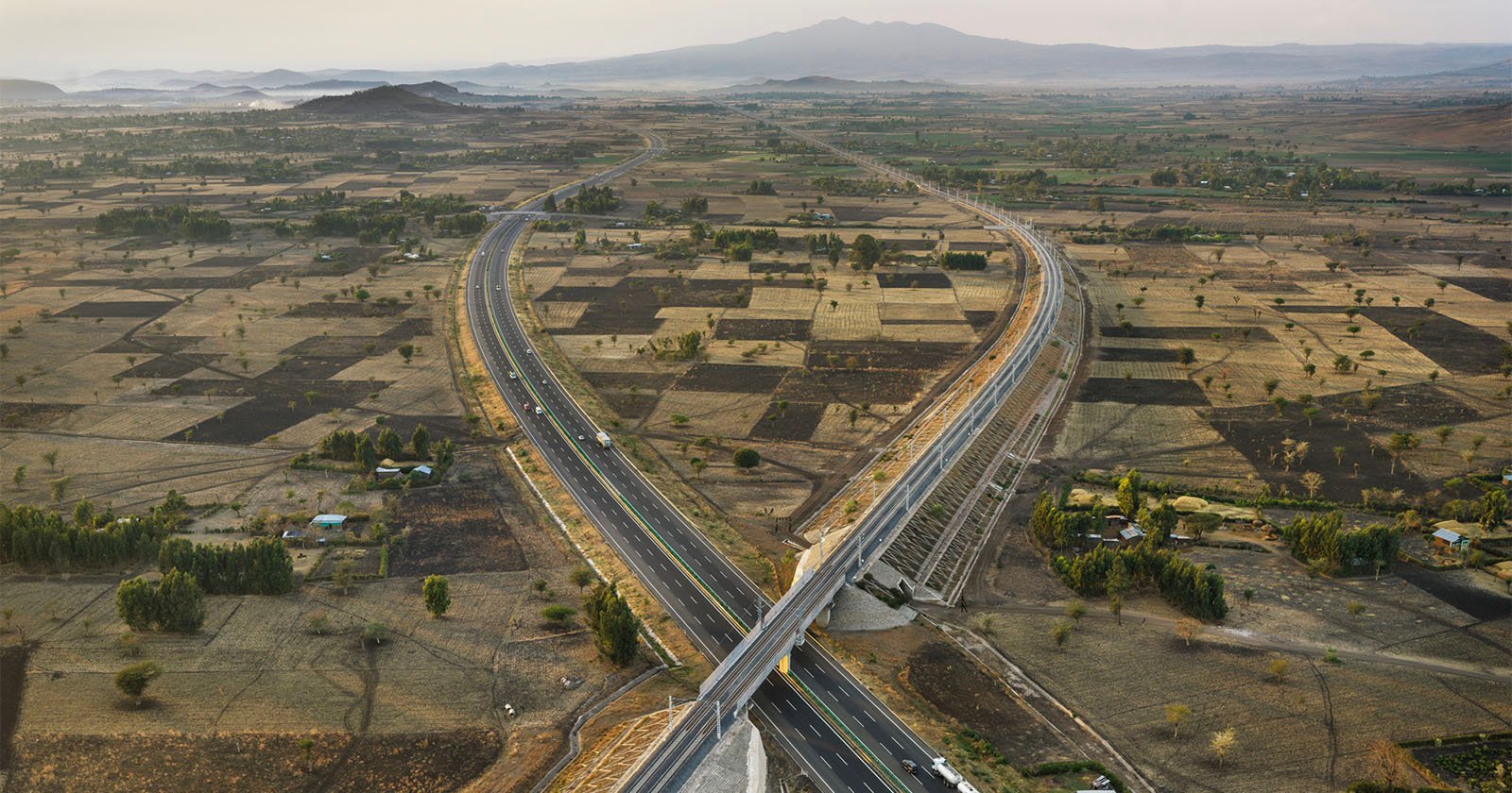 Aerial view of a modern highway interchange crossing over patchwork farmland, with cars on the roads and distant mountains under a hazy sky in the background.