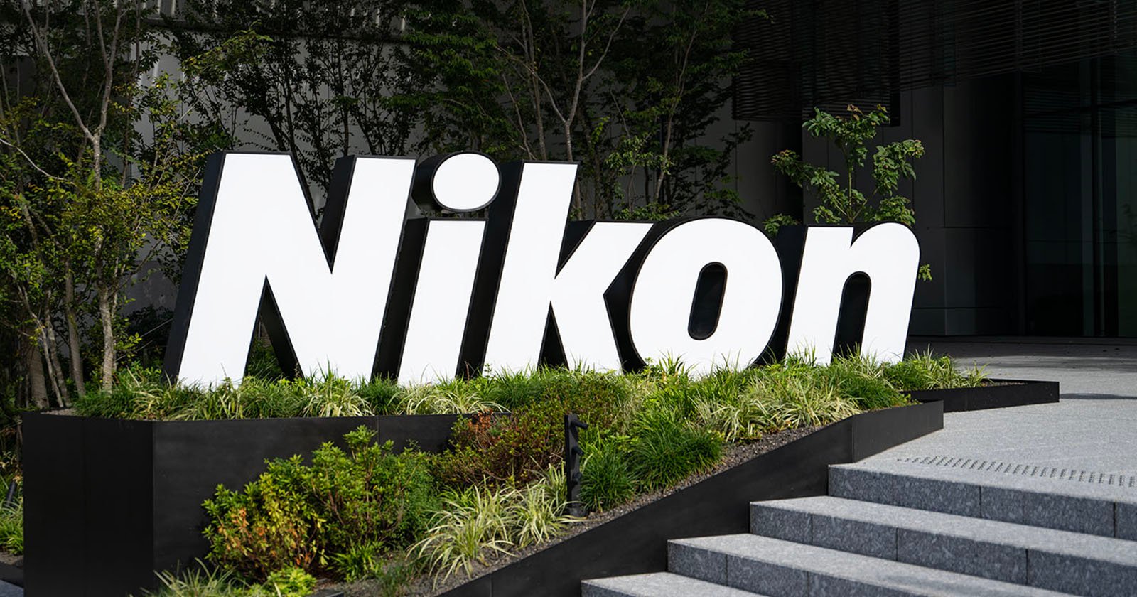 Large, white Nikon sign stands amid greenery outside a modern building, near concrete steps and surrounded by trees and bushes.