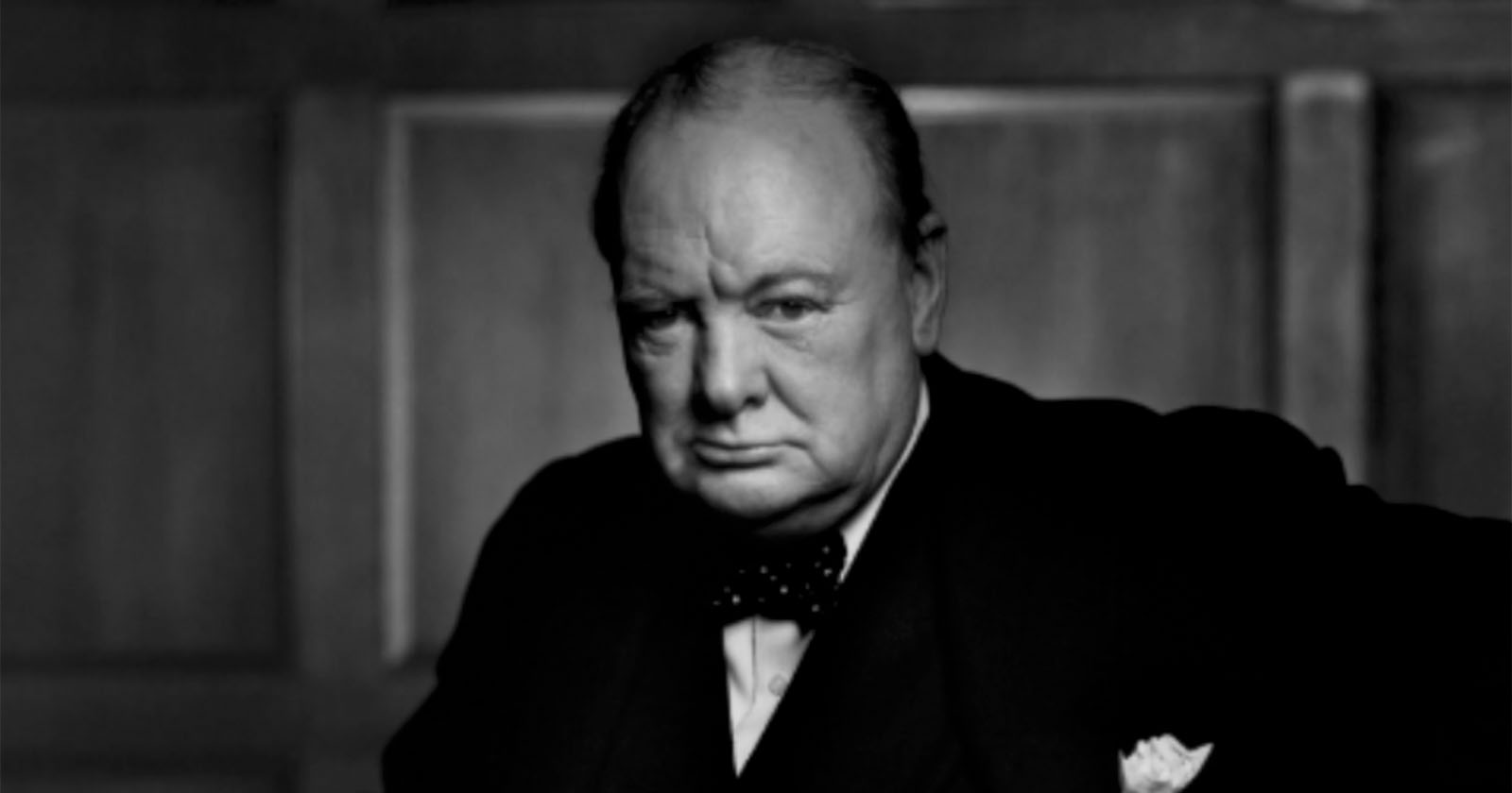 A monochrome photograph of an elderly man in formal attire, including a black suit, white shirt, and bow tie. He has a serious expression and is positioned against a wooden panel background.