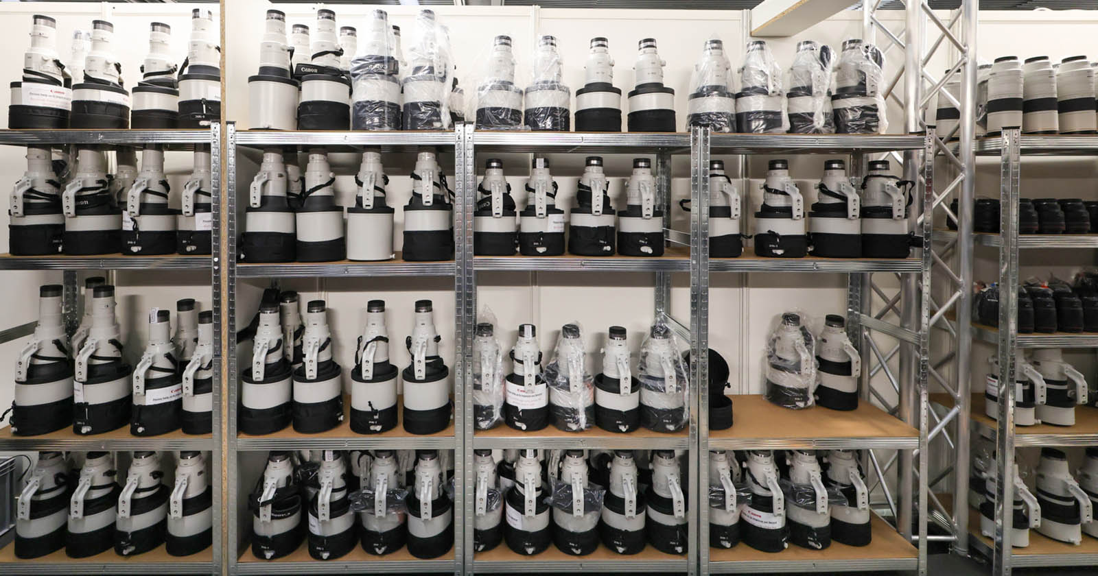 A storage room with metal shelves filled with numerous large camera lenses, many of which are wrapped in plastic. The lenses primarily have white and black color schemes and are neatly arranged in rows on the shelves.