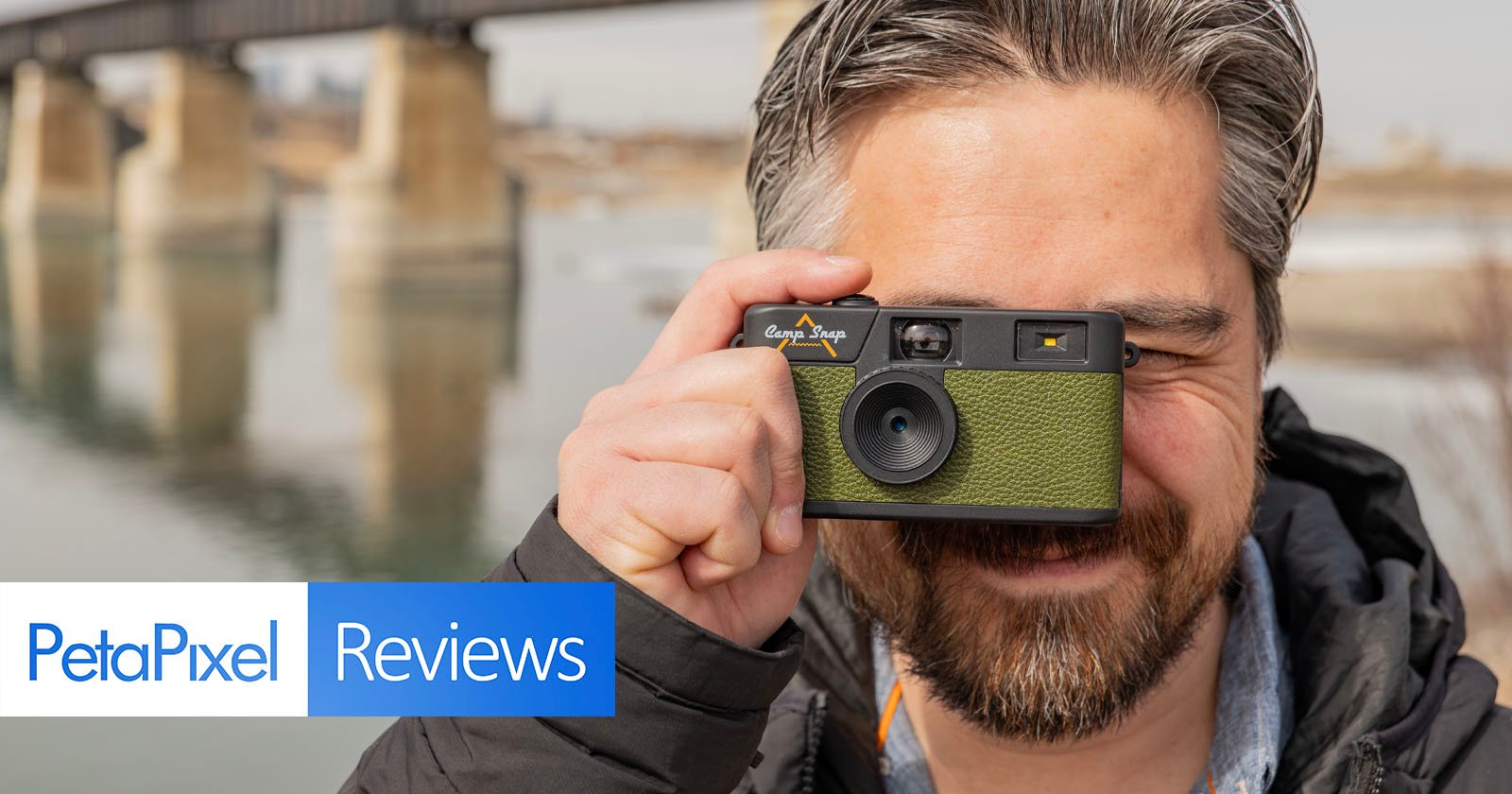 A person holding a green film camera up to their eye, smiling, with a bridge and water in the background. The image includes text reading PetaPixel Reviews at the bottom left corner.
