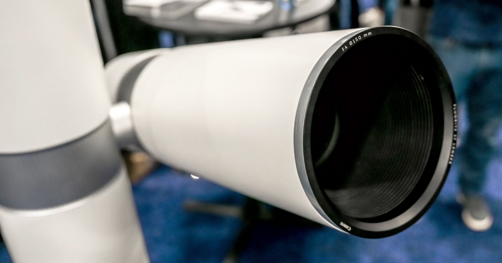 Close-up view of the lens end of a large, gray telescope with a black rim, displayed indoors on a blue carpeted floor with blurred background.