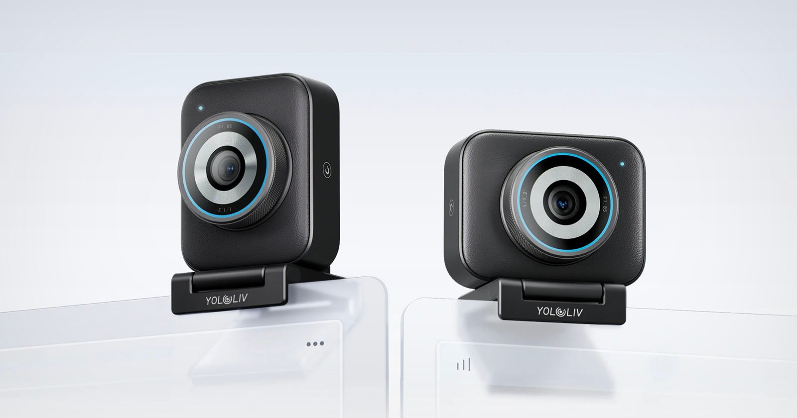 Two black rectangular webcams with circular blue accents are mounted on top of white computer monitors. The brand YoloLiv is visible on each webcam's base. The background is plain and light-colored.