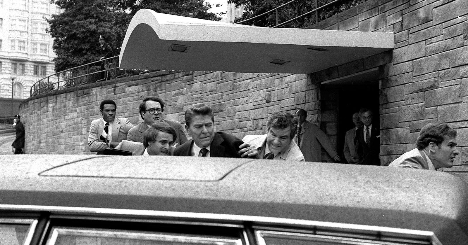 A black and white photo shows several men in suits rushing out of a building with a covered entrance. A prominent figure in the center is being assisted into a waiting car, as others appear to protect him. The scene is urgent and seems to involve security personnel.