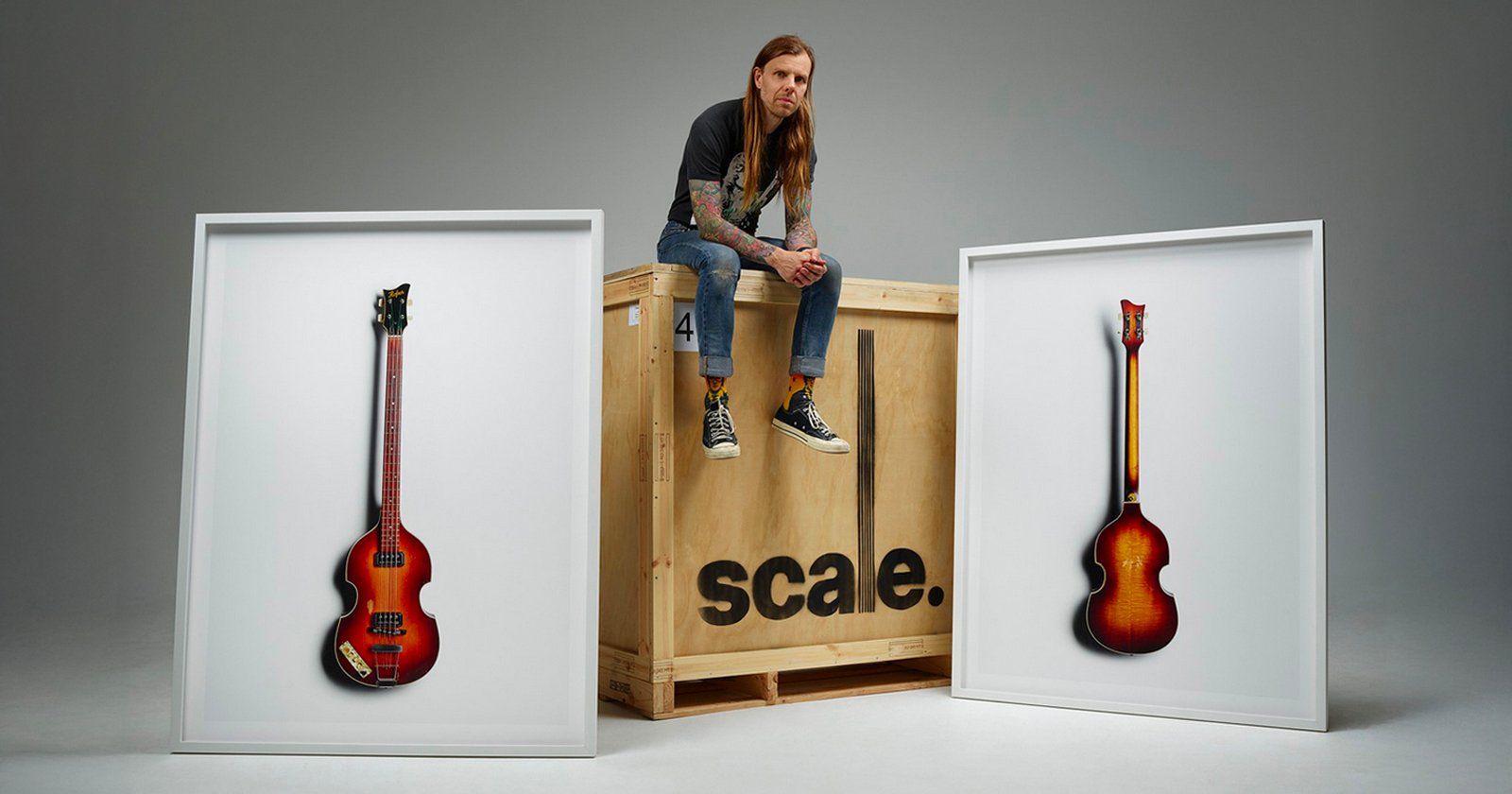 A man with long hair and tattoos sits on a large wooden crate labeled scale. Two framed photos of vintage guitars are displayed on either side of the crate against a plain gray background.