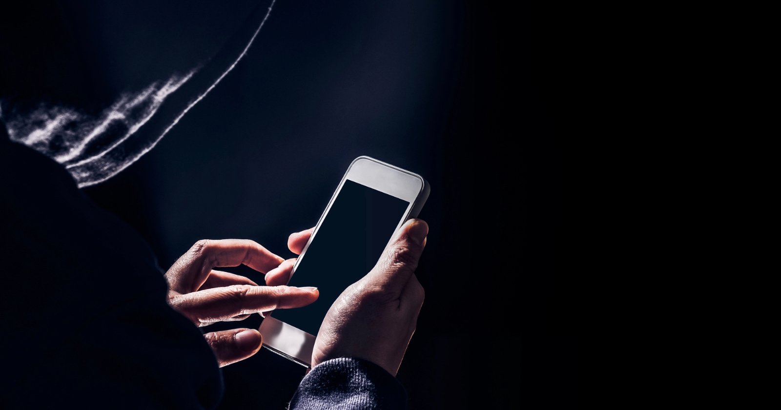 A person wearing a hoodie uses a smartphone in a dark setting, with their face obscured by shadows, creating a mysterious or secretive atmosphere.