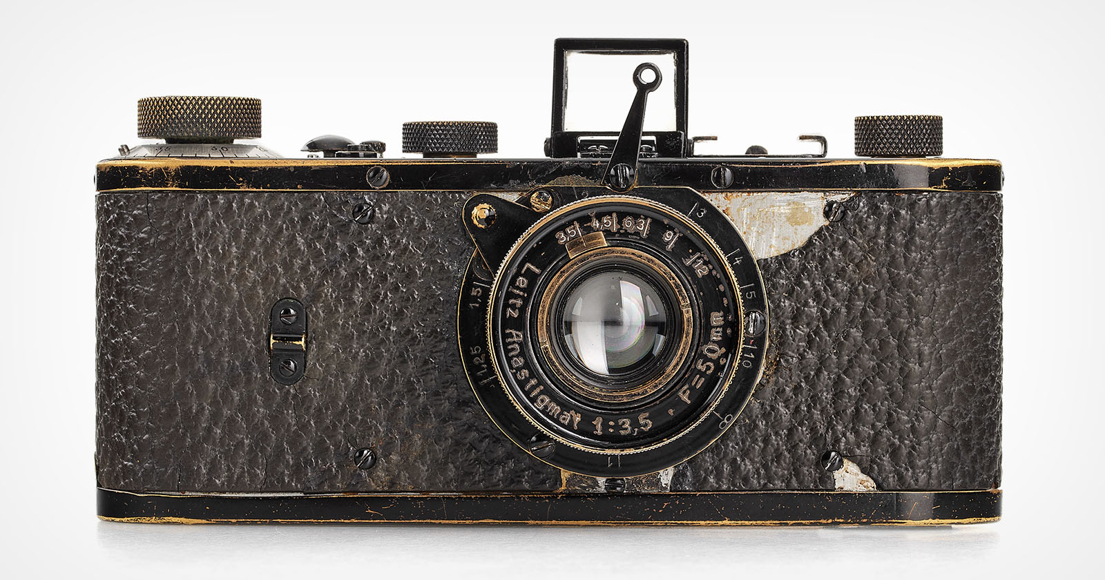 Vintage Leica camera with a worn, textured body and brass accents. The lens reads Leitz Anastigmat f=5cm 1:3.5 and shows signs of age. The camera has a classic, rectangular design with various dials and a viewfinder on top.