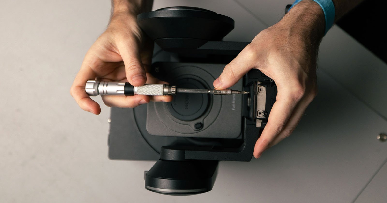 A person uses a screwdriver to repair or assemble a black camera device, viewed from above, with both hands visible holding the tool and device.