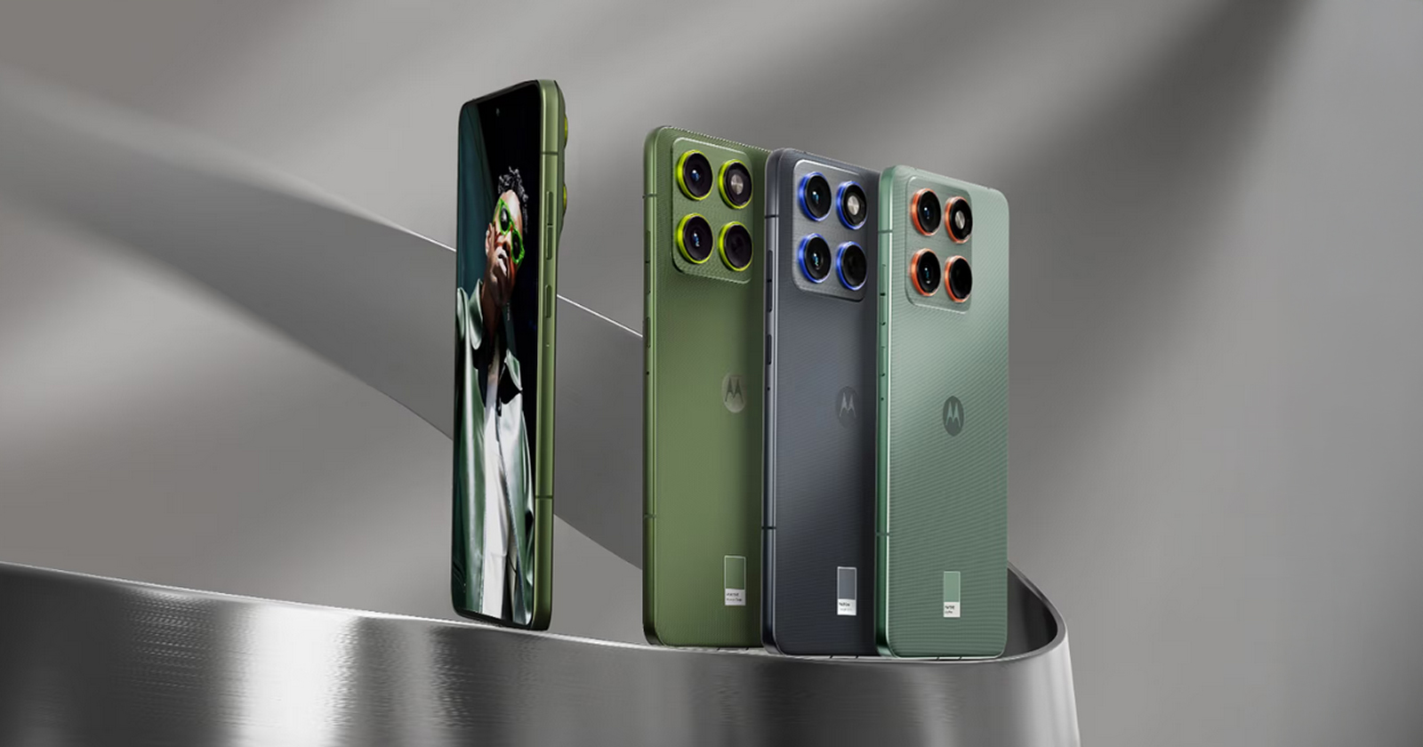 Four Motorola smartphones are displayed on a curved metallic surface; three show their stylish rear designs in green, blue, and greenish-gray, while one shows its front with a portrait on the screen.