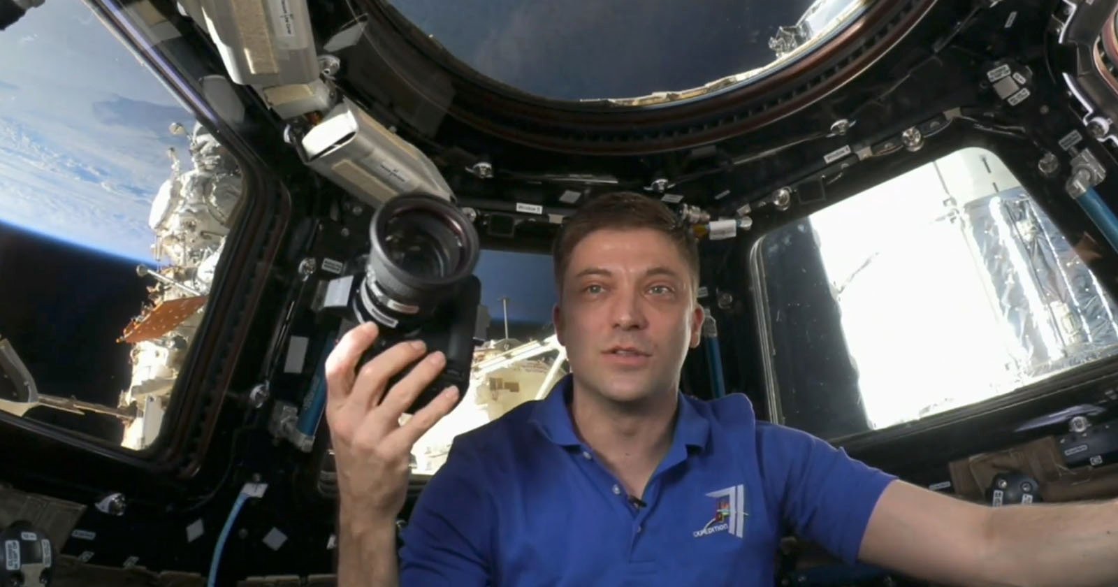 An astronaut inside a spacecraft holds a camera, with Earth visible through the windows in the background. He wears a blue polo shirt and is surrounded by various equipment and controls. The scene is well-lit, showcasing the interior details of the spacecraft.