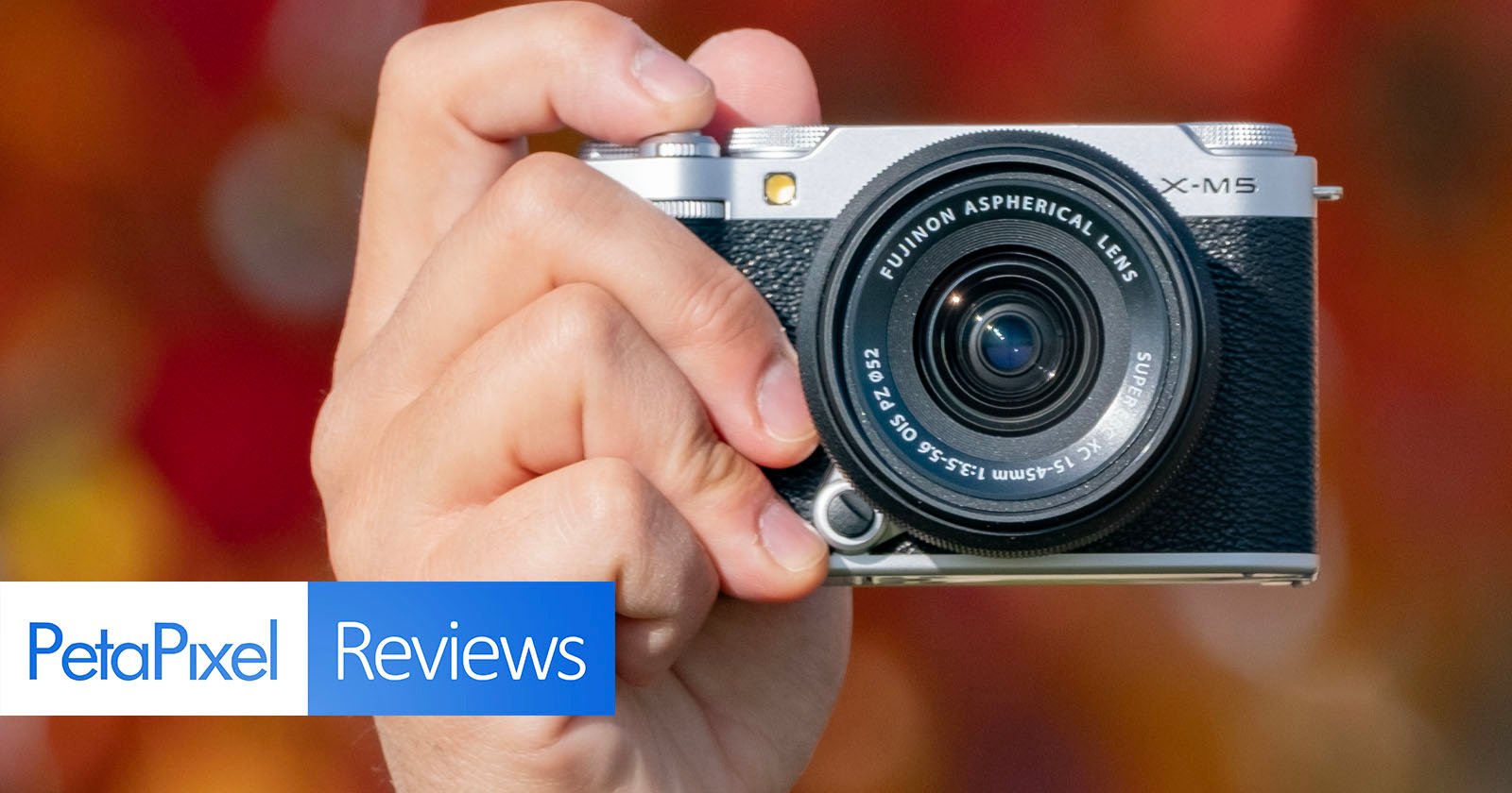 A hand holding a Fujifilm X-M5 camera with an aspherical lens. The background is blurred, showcasing autumn colors. Overlay text at the bottom reads PetaPixel Reviews.