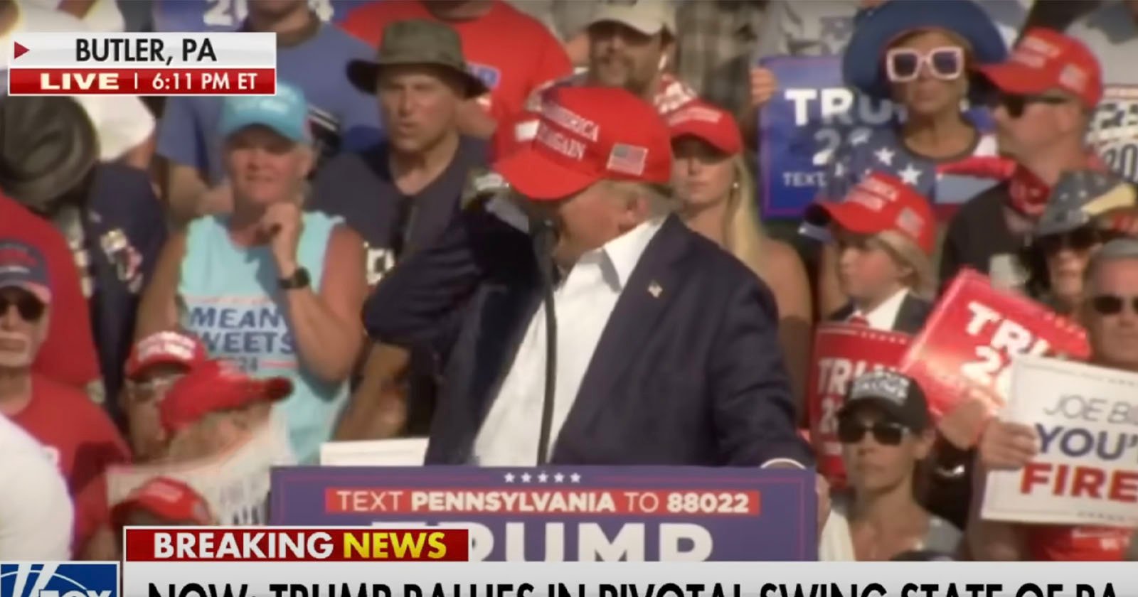 A crowd gathers around a podium with Trump signage at a rally in Butler, PA. Many attendees wear red caps and hold signs. A man at the podium, wearing a red cap, speaks into a microphone. News ticker below reads, BREAKING NEWS NOW: TRUMP RALLIES IN PIVOTAL SWING STATE OF PA.  .