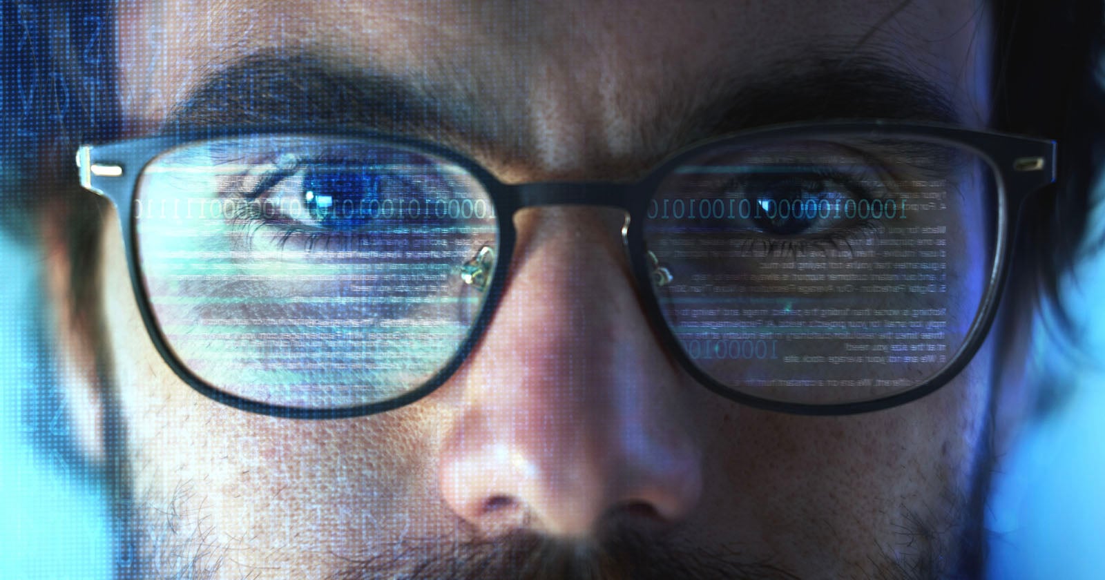 Close-up of a person's face with glasses reflecting computer code and binary numbers, suggesting intense focus on a digital screen, technology, or cybersecurity.