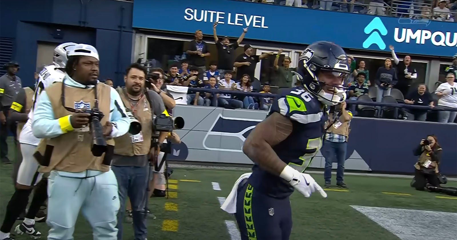 A Seattle Seahawks player runs along the sideline during a football game as photographers and fans watch from behind the barrier. The background shows excited fans and a Suite Level sign.