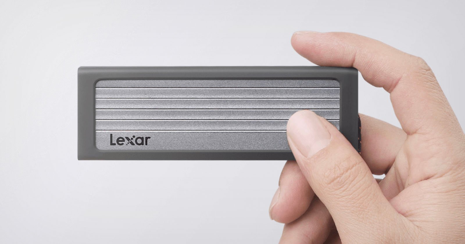 A hand holding a Lexar portable solid-state drive (SSD). The SSD is rectangular with a dark frame and a metallic center featuring horizontal ridges. The Lexar logo is visible on the bottom left corner of the SSD.