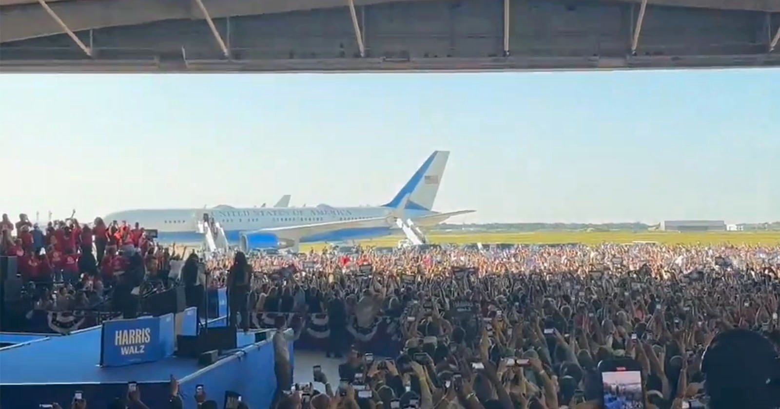 A large crowd gathers in an open hangar for a political rally. The stage displays a Harris Wally sign. Two large aircraft, including one with United States of America marked on it, are parked in the background against a clear sky. Many people are taking photos.