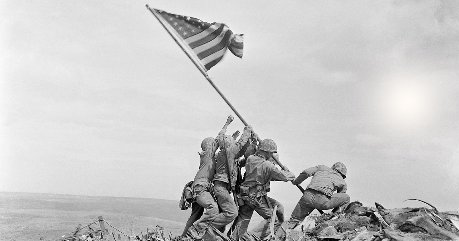 Five soldiers raising an American flag on a pole atop a rugged hill during a historical battle. The sky is clear, emphasizing the significance of the moment. The image captures a sense of teamwork and patriotism.