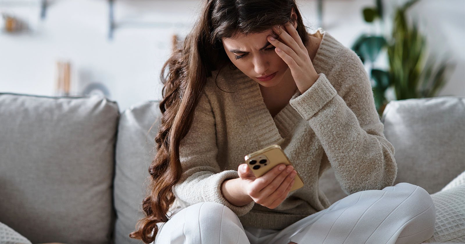 A woman with long brown hair sits on a couch, looking stressed as she holds a smartphone in one hand and rests her other hand on her forehead. She appears worried or upset.