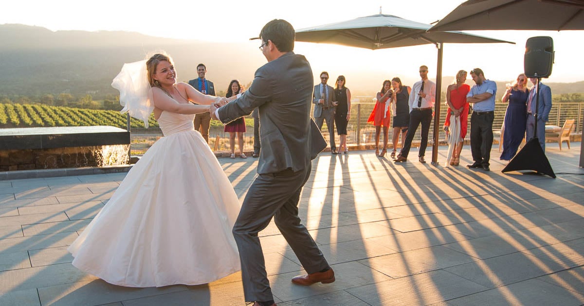 A bride in a white dress and veil and a groom in a gray suit joyfully dance at an outdoor wedding reception with vineyards in the background. Guests, some holding drinks, watch and smile under patio umbrellas as the sun sets.