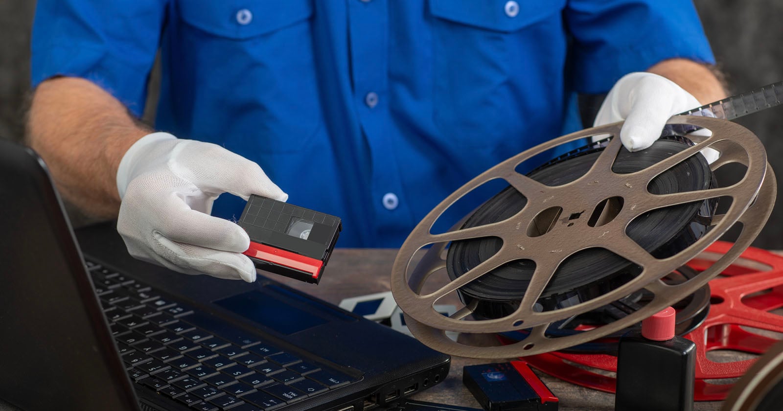 A person wearing white gloves holds a film reel in one hand and a VHS cassette in the other, working at a desk with a laptop, red film reels, and video equipment.