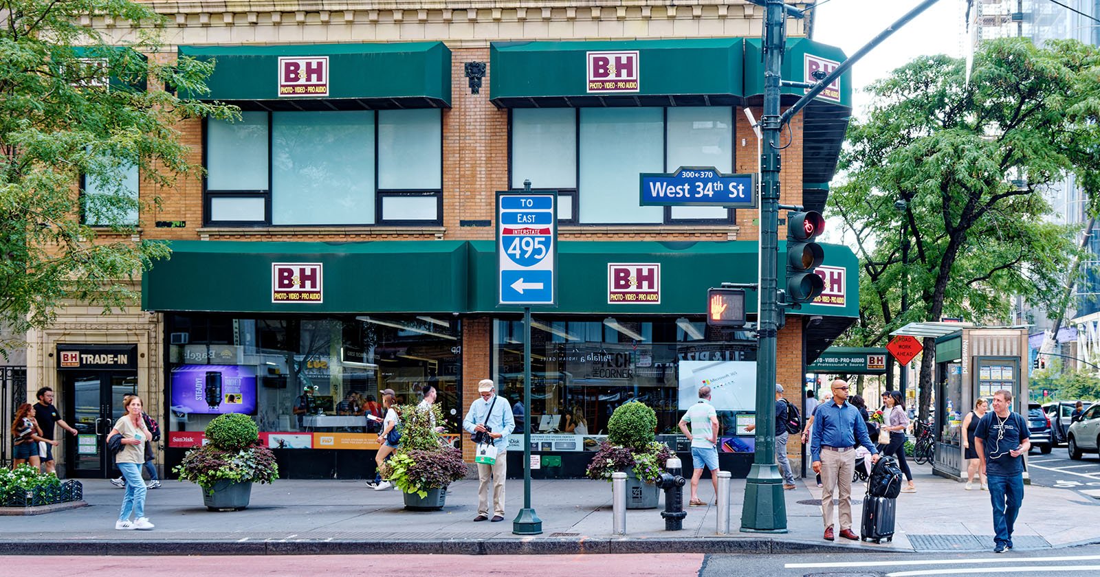 Street view of a busy storefront with B&H signage on awnings. People are walking on the sidewalk, and trees and potted plants decorate the scene. Traffic signs point to East 495/West 34th St. It's a bustling urban environment.