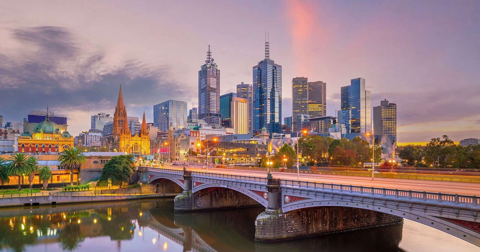 A cityscape of Melbourne at dusk, showing modern skyscrapers, historic buildings, streetlights, and a bridge over the Yarra River with reflections in the water.
