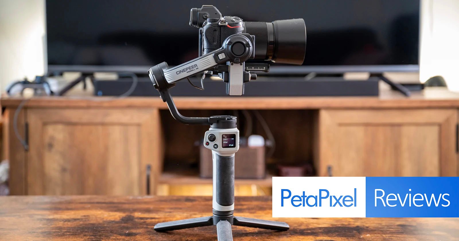 A professional camera is mounted on a stabilizing gimbal, which is placed on a wooden surface. In the background is a wooden console with a television. The photo has overlapping text that reads PetaPixel Reviews in the lower right corner.
