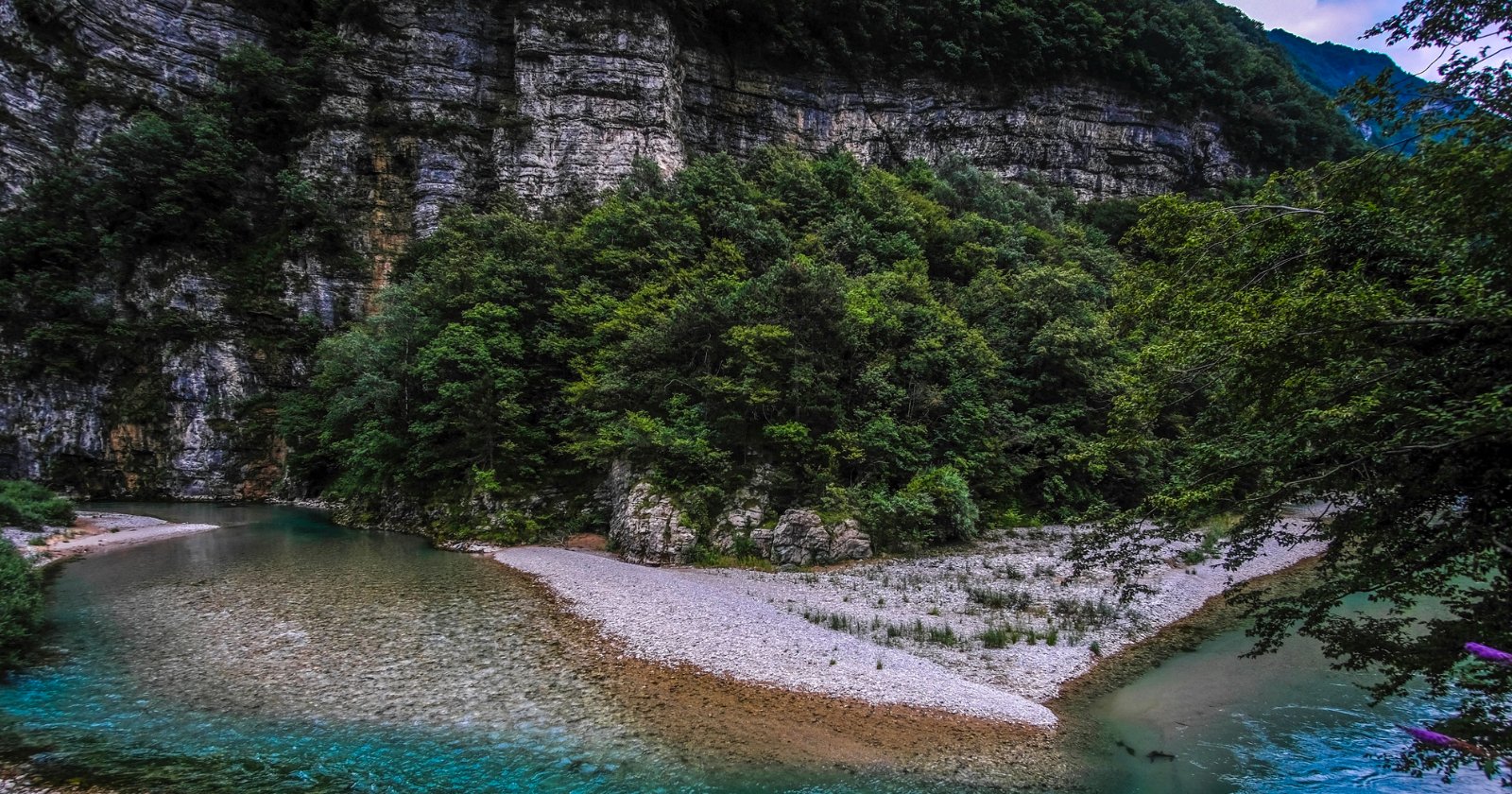 A clear river curves around a gravelly, tree-covered peninsula, with steep, rocky cliffs and dense green forest rising in the background under a cloudy sky.