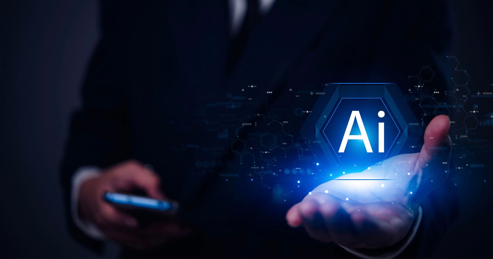 A person in a suit holds a glowing hexagonal icon with the letters AI above their open palm, representing artificial intelligence, while using a smartphone in the other hand. The background is dark and tech-themed.