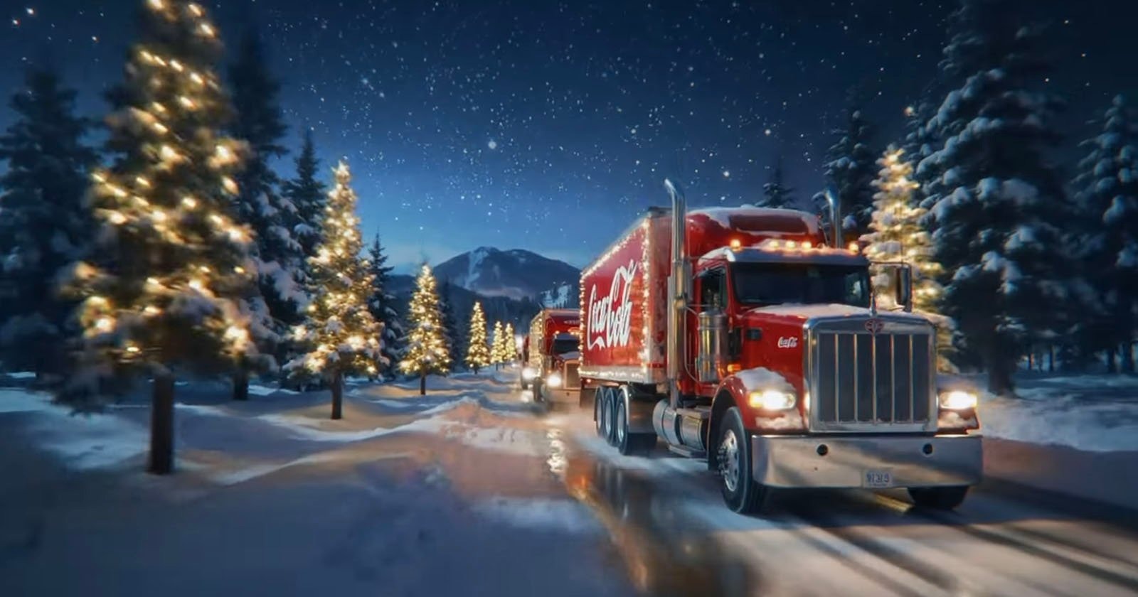 A red Coca-Cola truck drives down a snowy, tree-lined road at night, with holiday lights on the trees and a starry sky above. Mountains are visible in the background.