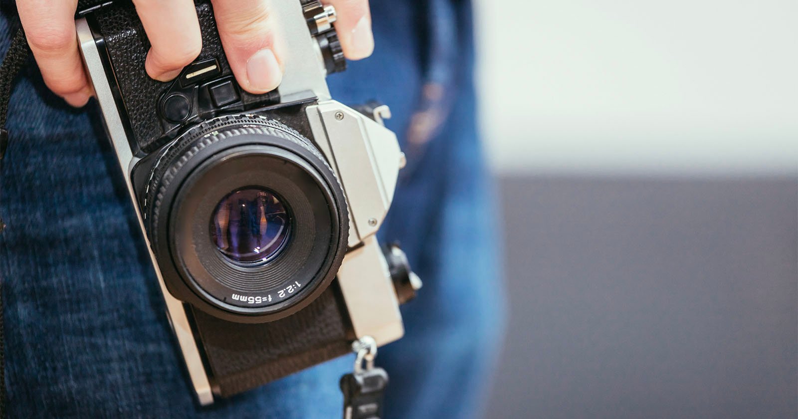 Close-up of a person holding a vintage camera with a large lens. The camera is partially visible, and the person is wearing blue jeans. The background is blurred, emphasizing the camera in focus.