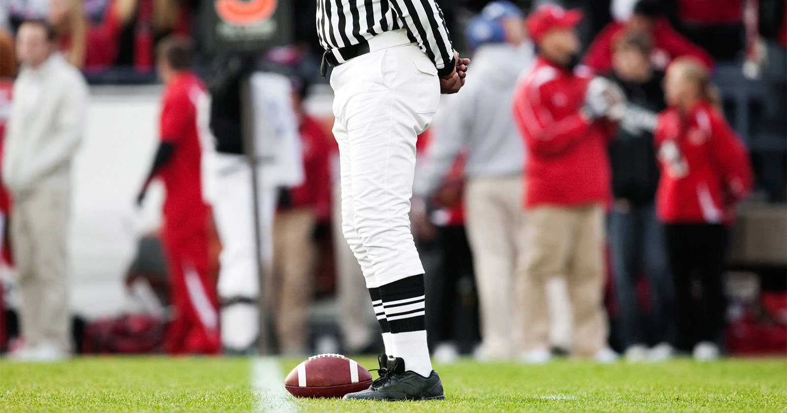 A referee in a black and white striped uniform stands on a football field next to a football. The background shows blurred figures in red, possibly team members, and spectators.
