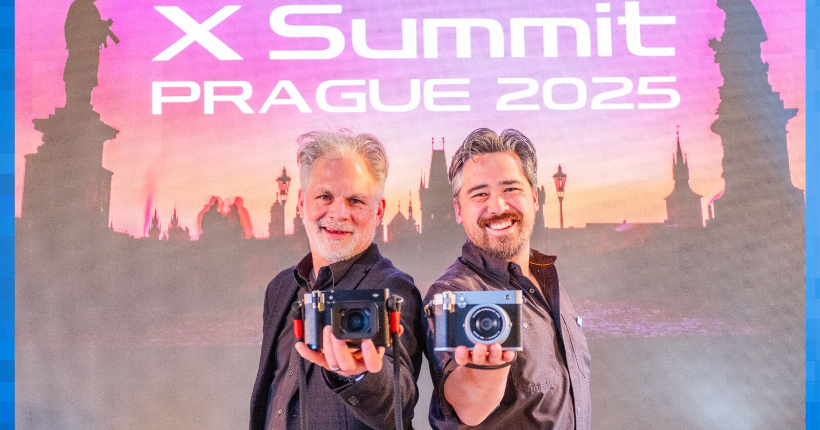 Two men standing in front of a backdrop reading X Summit Prague 2025 hold cameras towards the viewer. The backdrop shows a silhouette of a cityscape at sunset. Both men are smiling and dressed casually.