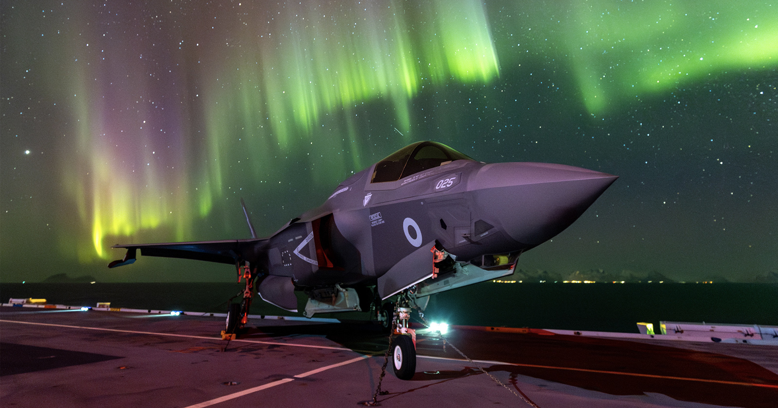 A fighter jet is parked on an aircraft carrier deck under a vivid display of the northern lights. The night sky is filled with colorful waves of green and yellow auroras, enhancing the dramatic scene.