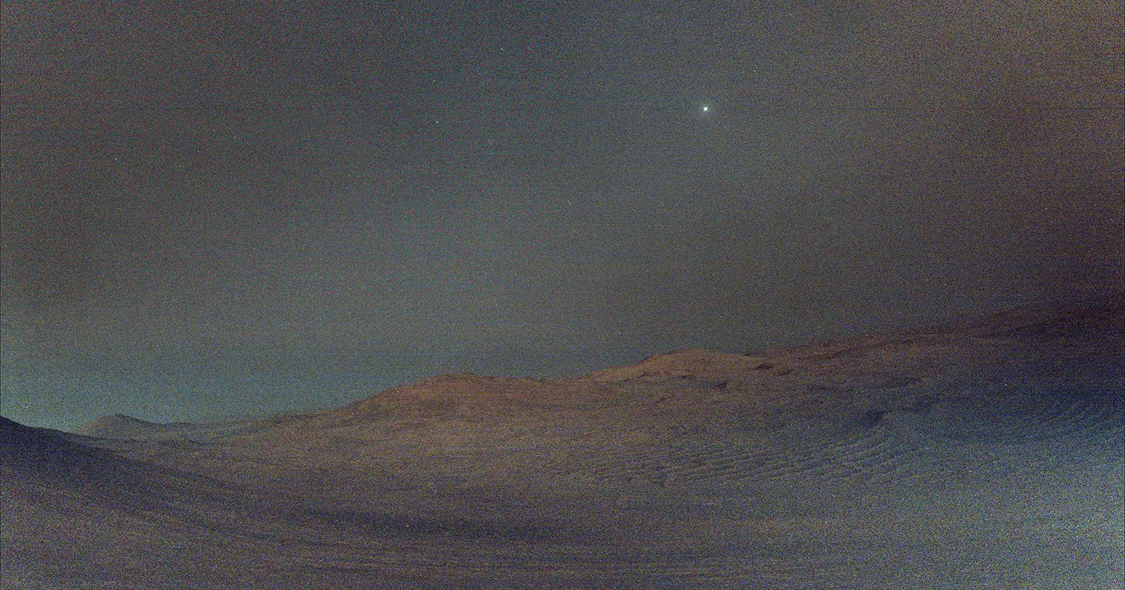 A dusty, rocky Martian landscape under a dark sky, with gentle hills in the distance. A bright white dot, representing the planet Venus, shines in the upper part of the night sky.