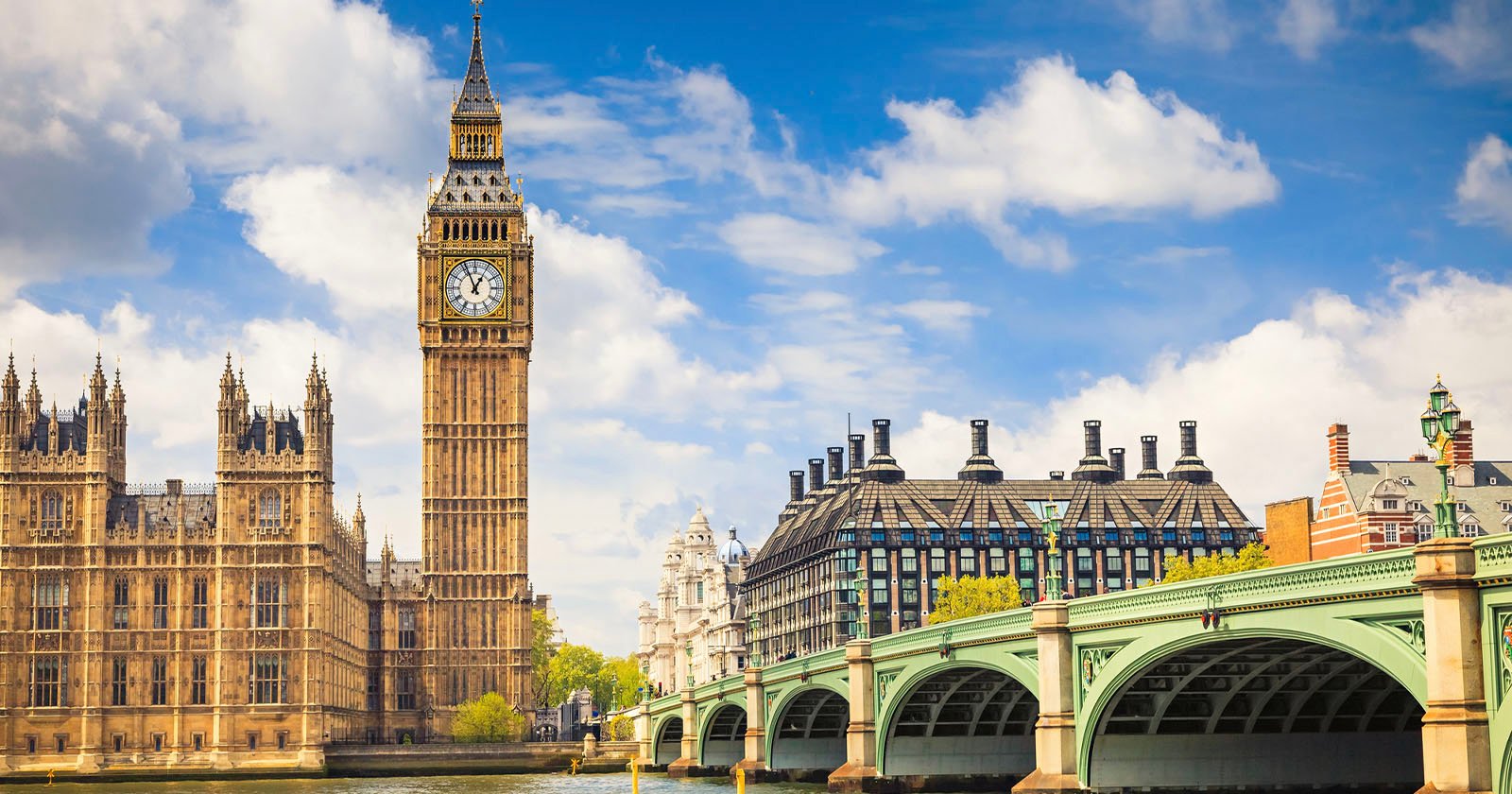 A view of Big Ben and the Houses of Parliament in London, with Westminster Bridge crossing the River Thames under a partly cloudy blue sky.