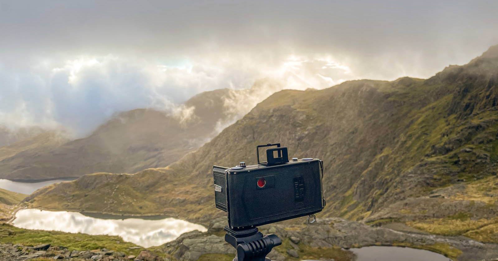 A vintage camera on a tripod faces a misty mountain landscape with rolling hills, rocky terrain, and reflective lakes under a dramatic cloudy sky.