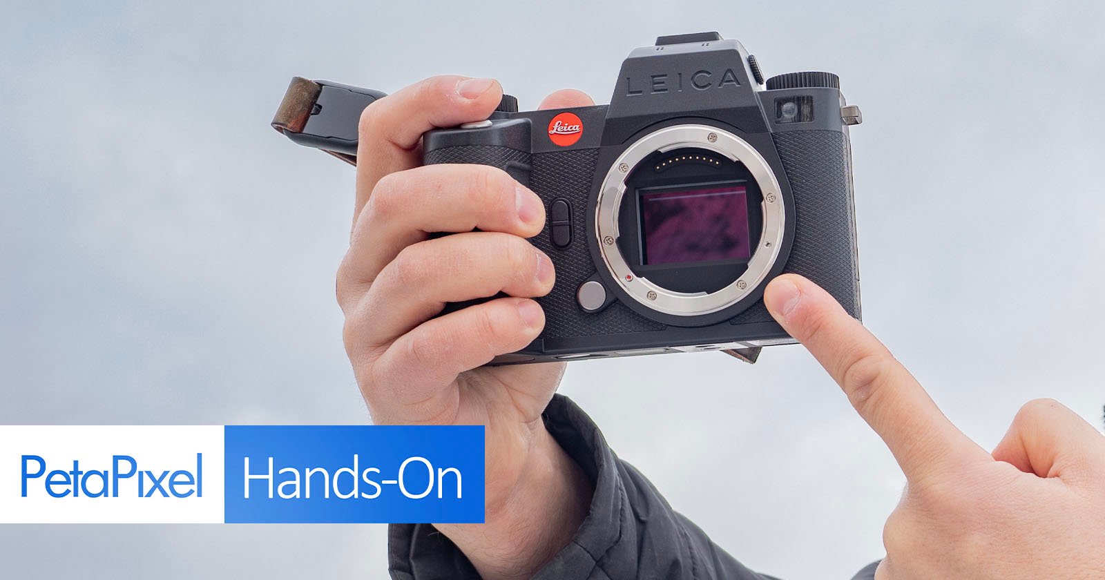 A person holding a Leica camera, pointing at the sensor with one finger. The image includes the PetaPixel logo and the words Hands-On at the bottom left corner. The camera is shown against a blurred gray background.