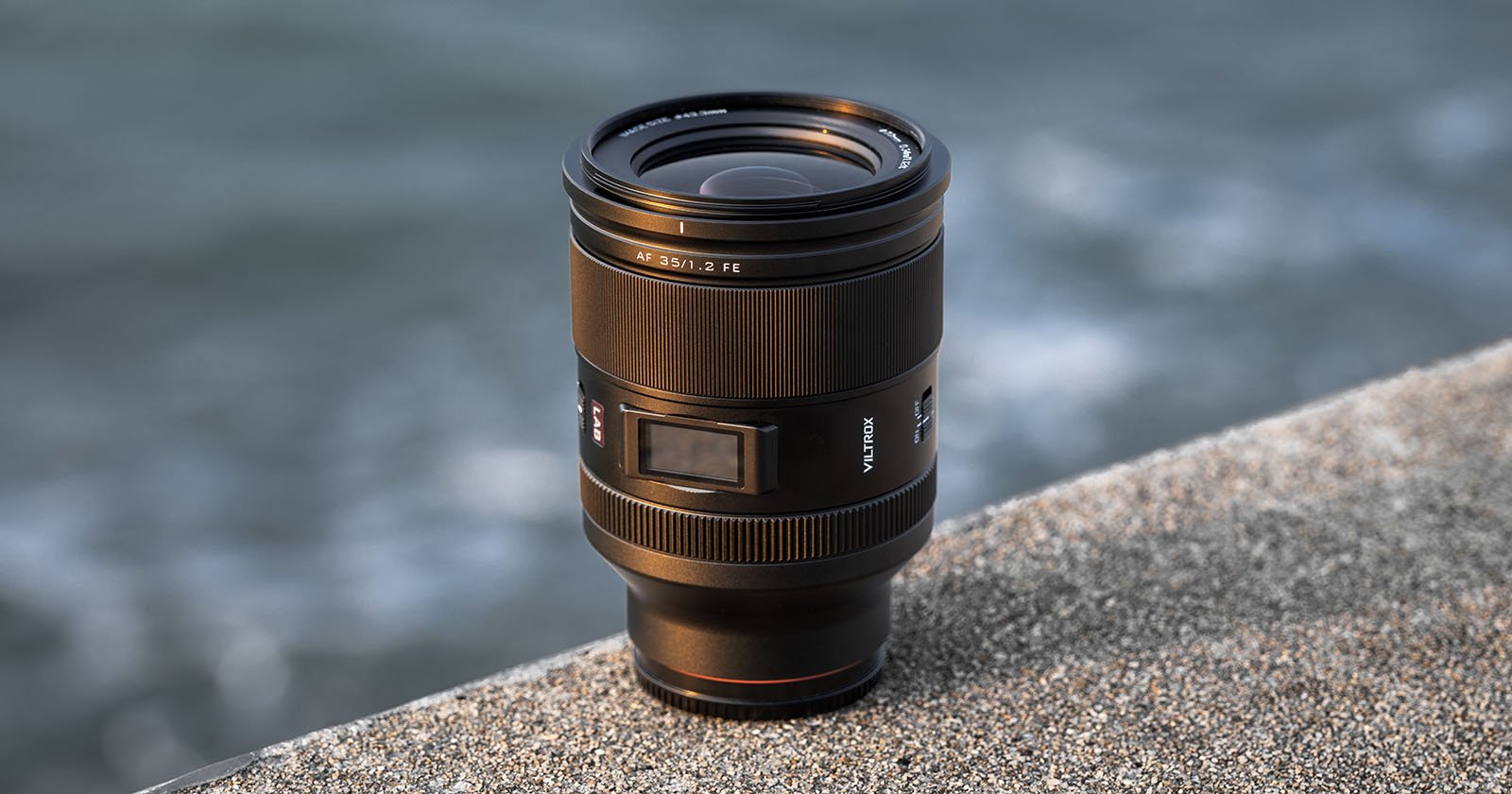 A camera lens stands upright on a rough concrete surface near a body of water, with the background softly blurred.