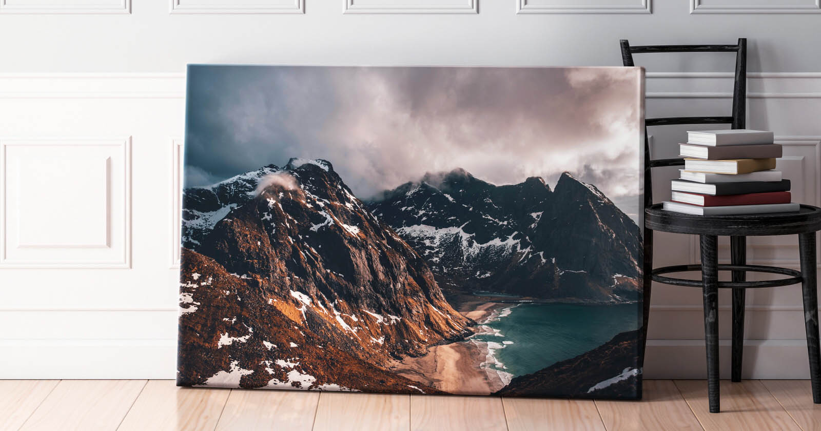 A large framed photograph of a dramatic mountainous landscape with snow-covered peaks and a coastline with a sandy beach leaned against a white-paneled wall. It’s positioned beside a wooden chair stacked with several books.