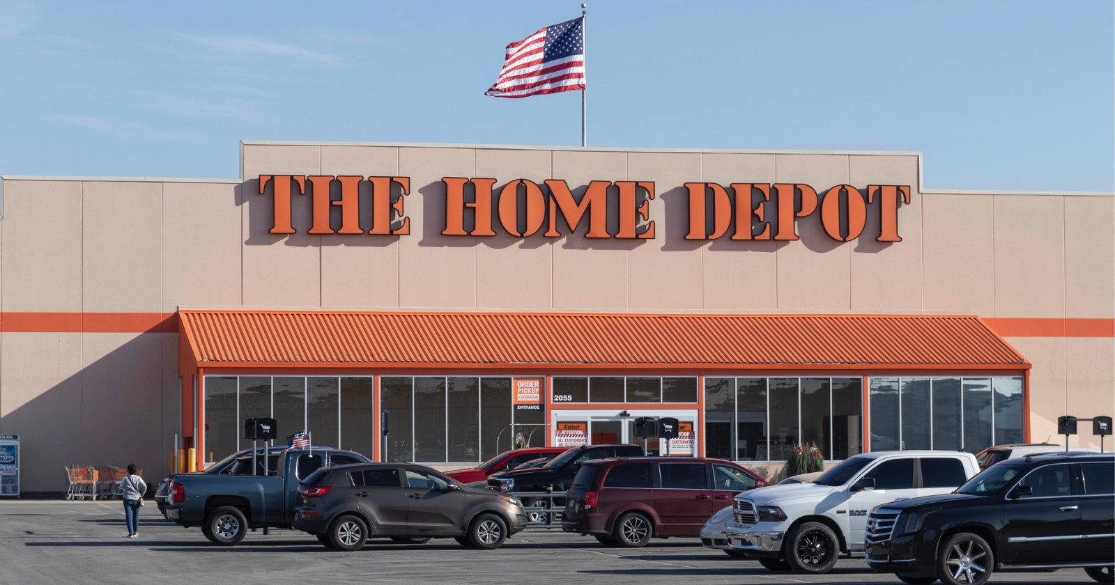 The Home Depot store exterior with an American flag on the roof and several cars parked in front of the entrance on a sunny day.