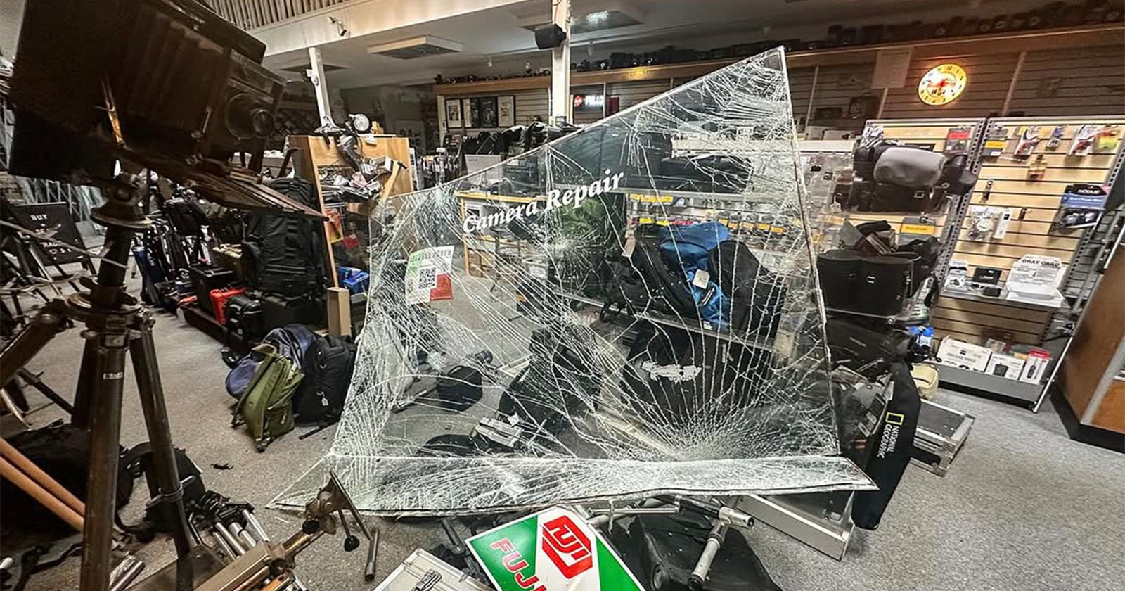 A camera repair shop interior with a large glass window shattered into many cracks. Various camera equipment and bags are visible in the background. Lighting equipment is positioned on the left side.
