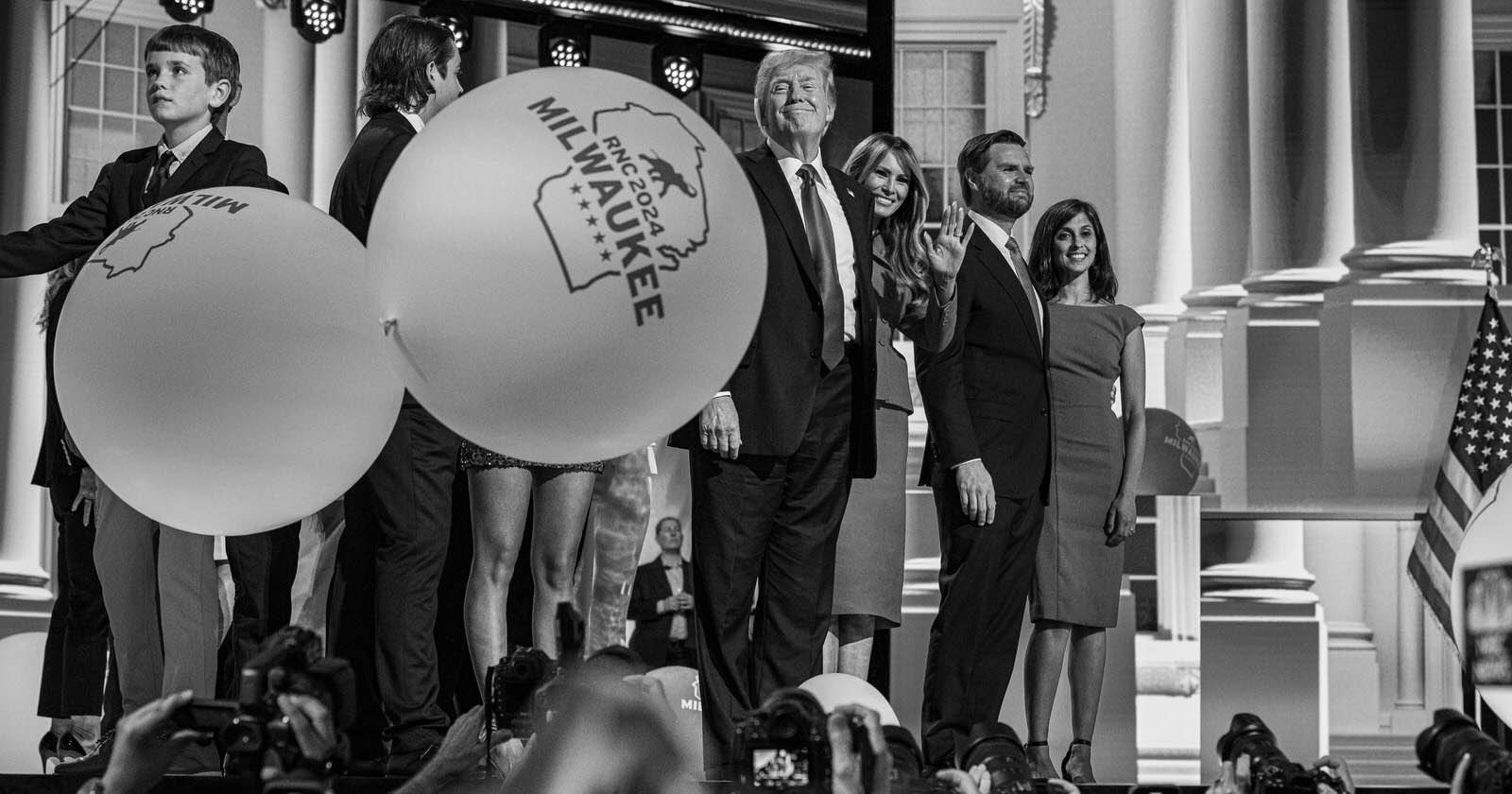 A group of people stands on stage in front of a backdrop resembling the White House. Two large balloons with MILWAUKEE and a star are near the left side. Cameras and onlookers are seen in the foreground capturing the scene. The image is in black and white.