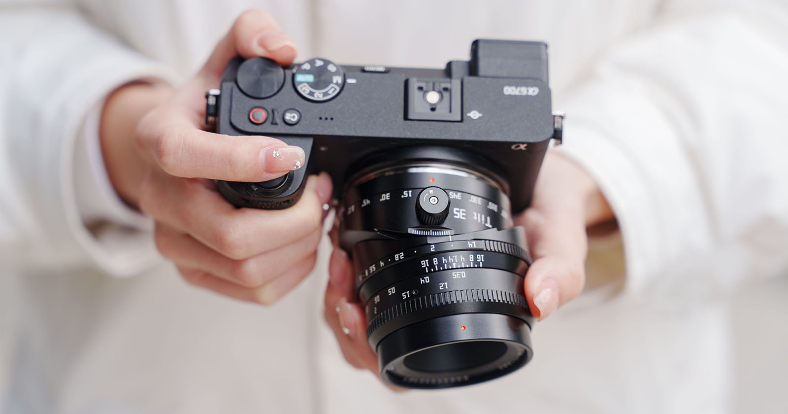 A person in a white jacket holds a black digital camera with both hands, adjusting the lens and dials. The camera and the person’s hands are in sharp focus.