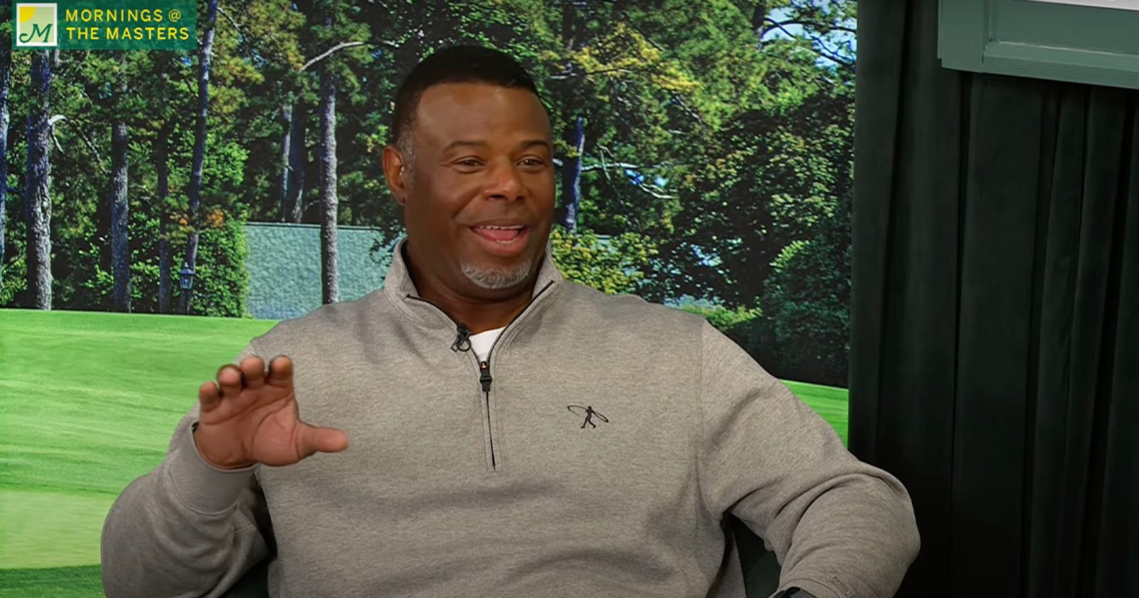 A man in a grey sweater is smiling and gesturing with his hand while sitting. The background shows a forest scene. The top left corner displays the text MORNINGS @ THE MASTERS with a green and yellow logo.